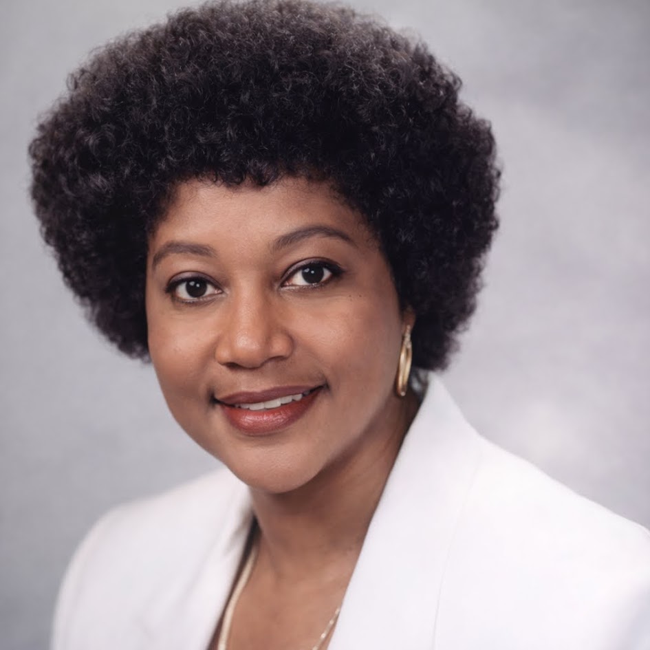 Close-up portrait of a woman with natural curly hair, wearing a white blazer and gold earrings, smiling at the camera against a gray background.